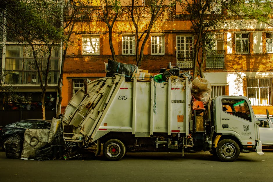 A waste collection truck parked on a residential street in Putney, with its rear loaded with large bags of rubbish, cardboard boxes, and discarded items. The truck is white with red and black markings, positioned in front of a multi-storey brick building with tall windows and metal railings. The street is lined with trees casting shadows on the building, and a black car is parked nearby. The scene depicts the process of waste removal, relevant to bulky waste disposal and interior cleaning tasks, emphasizing the importance of proper waste management before or after deep cleaning services offered by Cleaners SW15.