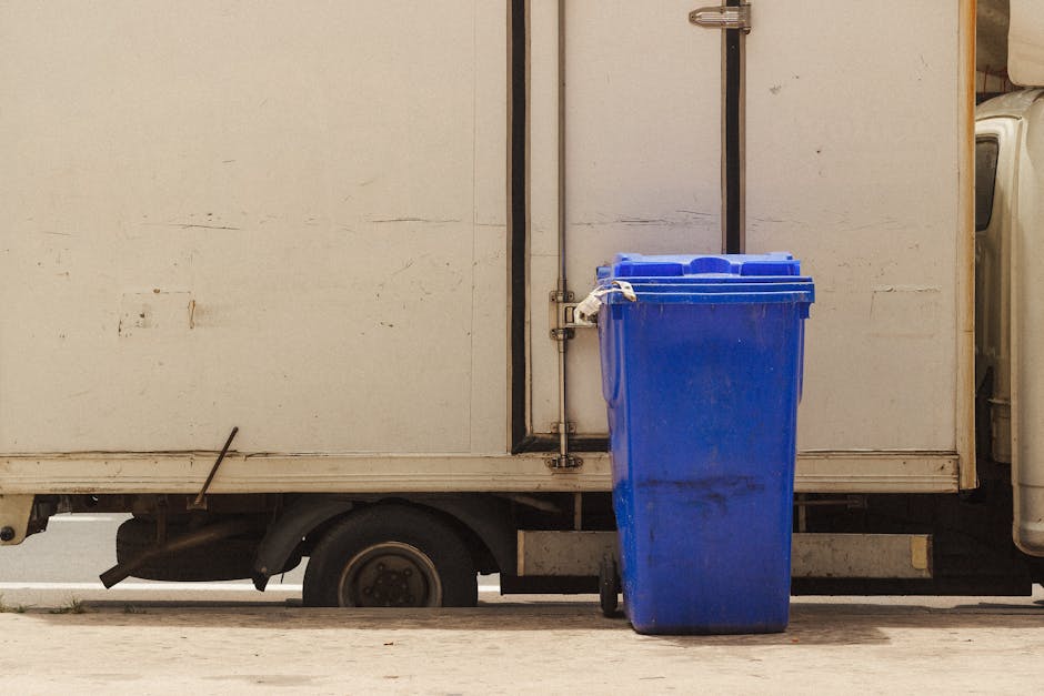 A blue recycling bin positioned on a paved surface beside a white commercial vehicle or food truck, with a closed lid and slightly darkened markings stains on its surface. The vehicle has visible signs of dirt and wear, with a metal pipe running vertically along its side, and a partially visible washing machine or appliance to the right. The scene is lit with natural daylight, highlighting the surfaces' textures and cleanliness levels, indicating a setting where surface cleaning or waste disposal processes are involved. The setting emphasizes the importance of sanitation and proper waste management in commercial or residential cleaning contexts, reflecting services provided by Cleaners SW15, particularly in relation to bulky waste removal and deep cleaning.