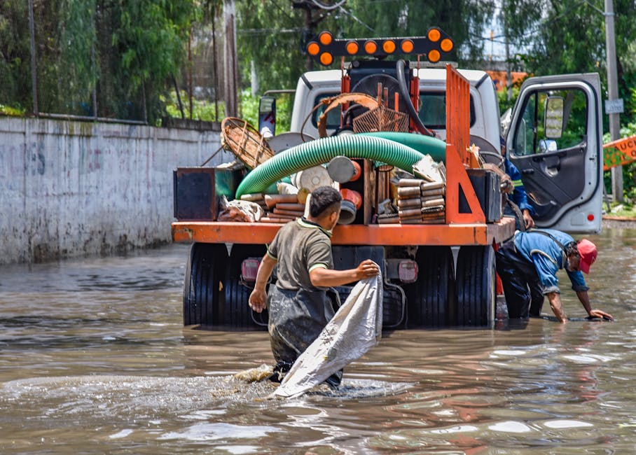 A residential street scene showing multiple workers in yellow and orange raincoats and protective gear engaged in emergency flood cleaning. The street surface is wet and partially flooded, with visibl