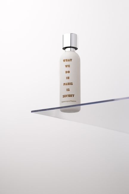 A white cylindrical bottle with a metallic silver cap, featuring brown text that reads 'WHAT WE DO IN PARIS IS SECRET' by Dominique Ropion, positioned on a transparent glass shelf against a plain white wall. The surface of the bottle appears clean and new, with no visible dust or smudges, indicative of careful surface cleaning and sanitisation. The lighting highlights the smooth, glossy finish of the bottle, and the shelf is free of clutter or fingerprints, emphasizing a tidy and hygienic environment consistent with professional cleaning standards. This image reflects cleanliness and attention to detail in a domestic or commercial setting, suitable for content related to surface cleaning, deep cleaning, or hygiene maintenance, and naturally aligning with the topic of end-of-tenancy cleaning costs covered by Cleaners SW15.