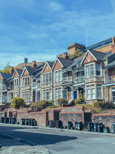 A row of Victorian-style terraced houses with brick and white decorative trim, featuring large bay windows and ornate iron balconies. The buildings are set on a slightly elevated street level with a brick retaining wall and steps leading up to each property. The pavement in front is lined with black wheelie bins and small landscaped areas with shrubs and plants. Bright sunlight illuminates the scene, highlighting the clean, well-maintained appearance of the facades and surroundings, reflecting thorough surface cleaning and maintenance performed by professionals such as those at Cleaners SW15, as discussed in the End-of-Tenancy Cleaning Cost in SW15 page.
