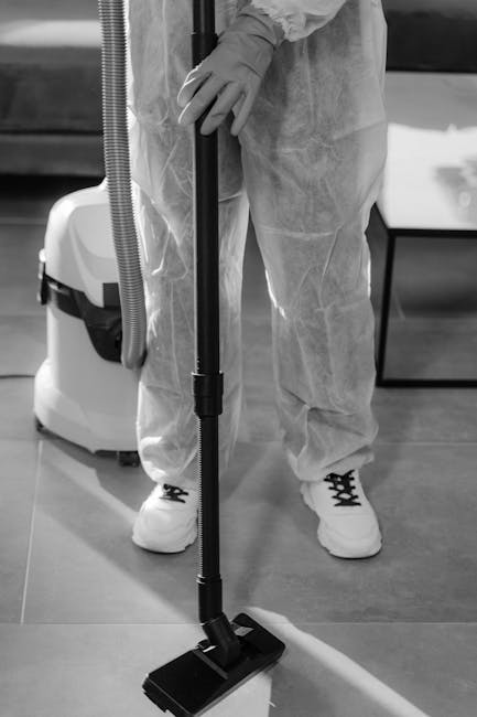 A professional cleaner from Cleaners SW15 is using a vacuum floor cleaning machine on a smooth, hard surface floor in an indoor setting. The cleaner is dressed in protective clothing, including dispos