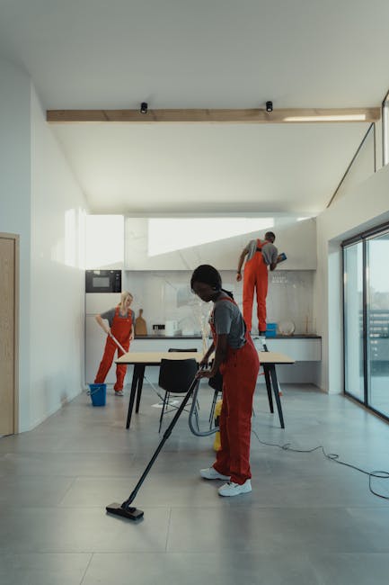A professional cleaner from Cleaners SW15 is using a vacuum floor cleaning machine on a smooth, hard surface floor in an indoor setting. The cleaner is dressed in protective clothing, including dispos
