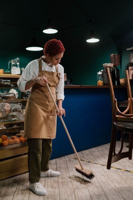 A person with curly red hair wearing a beige apron over a white shirt and dark pants is sweeping the wooden floor of a café with a long-handled broom. The café has a dark teal wall and a blue counter with glass display cases containing baked goods and fruits. Overhead, three round ceiling lights provide illumination, creating a well-lit environment. To the right, several wooden chairs are stacked upside down on a table, indicating a cleaning or closing time. The floor surface appears clean and dust-free, reflecting the café's hygiene standards. The presence of various cleaning tools suggests active maintenance of the venue, aligning with professional cleaning practices promoted by Cleaners SW15 on their Putney Cafe Cleaning Compliance page, which emphasizes proper sanitation and upkeep of commercial spaces.