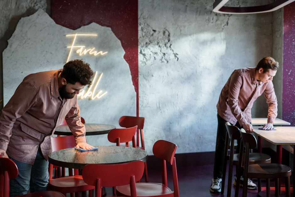 Two men in a modern cafe with exposed concrete walls, wooden tables, and red and black chairs, are engaged in surface cleaning and sanitisation. One man is wiping a round tabletop with a cloth, ensuring its cleanliness, while the other is disinfecting a rectangular table, both wearing casual brown shirts. The cafe features a neon sign on the wall that reads 'Food & Drink' in cursive, providing contemporary ambient lighting. The surfaces, including the tables and chairs, appear tidy and dust-free, with the wooden tabletops showing a slight sheen indicating recent cleaning. Natural light illuminates the space, highlighting the textures of the materials such as the smooth wood, concrete walls, and ceramic tiles. Cleaners SW15 specializes in domestic cleaning and deep cleaning services, ensuring hygienic standards are maintained for hospitality environments like this one, aligning with compliance requirements for cafes in Putney, SW15.