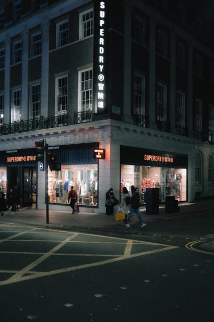 Nighttime exterior view of a multi-story retail building on Putney High Street, featuring illuminated signage for 'Superdry' and a vertical sign for 'Superdry' in English and Chinese characters. The ground floor has large glass storefront windows revealing the interior, with shoppers walking past the entrance. The street in front has marked crosswalks and traffic lights, with pedestrians crossing and waiting. The building's façade is made of brick and has white window frames, and there is subdued street lighting providing visibility. Cleaners SW15 specializes in surface cleaning and maintenance to ensure retail spaces remain hygienic and inviting, supporting the appearance of shops like Superdry during both daytime and evening hours.