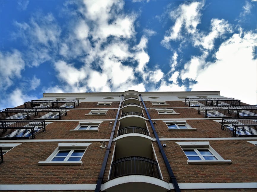 The image captures the exterior view of a multi-story residential building with a brick facade. The building features several small balconies with black metal railings, evenly spaced along the curved central portion. White-framed windows are visible on each floor, set into the brick walls. The sky above is bright blue with scattered white clouds, and the sunlight highlights the clean and well-maintained appearance of the building’s surface. The perspective is from ground level looking upward, emphasizing the height of the structure under natural daylight. This structural and cleanliness focus aligns with the services offered by Cleaners SW15 in surface cleaning and deep cleaning for residential properties like Riverside Flats by Putney Embankment, SW15.