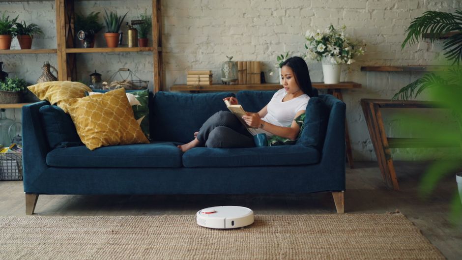 A woman is seated on a dark blue fabric sofa in a living room, holding a tablet and relaxing. The sofa has two yellow and one white decorative cushions. In front of the sofa, a round robotic vacuum cleaner is on a beige woven rug, indicating recent cleaning activity. The room features a white brick wall with open wooden shelving displaying potted plants, books, and decorative items, alongside a side table with a white vase of flowers and a stack of books. The space is well-lit with natural light, and there are various houseplants visible, creating a cozy and tidy atmosphere. This setting exemplifies professional domestic cleaning and surface maintenance, aligning with services offered by Cleaners SW15, as highlighted on the page about same-day cleaning in Putney.