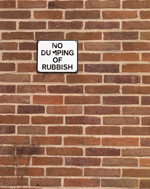 A close-up view of a red brick external wall featuring a small rectangular sign with a white background and black text that reads 'NO DUMPING OF RUBBISH'. The bricks are uniformly laid with mortar joints in shades of light gray, creating a consistent pattern. The lighting is natural, illuminating the brickwork evenly. The sign is mounted towards the upper left section of the wall, serving as a clear message to maintain cleanliness and hygiene standards in the area, which aligns with the themes of surface cleaning and maintenance promoted by Cleaners SW15 for residential and commercial spaces near Putney SW15. The overall scene emphasizes the importance of proper waste management to ensure a tidy environment.