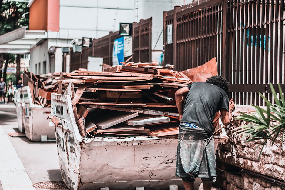 A man with curly hair wearing a black t-shirt and plaid shorts stands beside a large white skip filled with broken roof tiles and debris on a city sidewalk, with a brown metal fence and colored building facades in the background. The scene is outdoors during daylight, with shadows cast on the ground. The skip appears to be used for construction or renovation waste, and the area looks organized for waste removal, with a few other skips visible further along the sidewalk. Cleaners SW15 provides surface cleaning and waste management services, including deep cleaning and sanitisation, to assist with property maintenance and debris removal in residential or commercial settings.