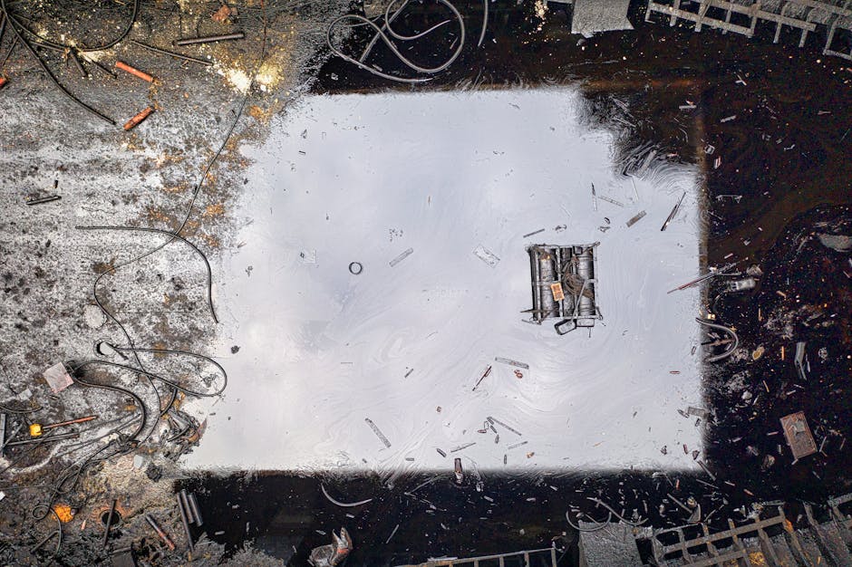 A square excavation site with a partially removed concrete surface, revealing an underlying layer of asphalt. The edges of the hole are rough and uneven, with scattered debris including wires, small t